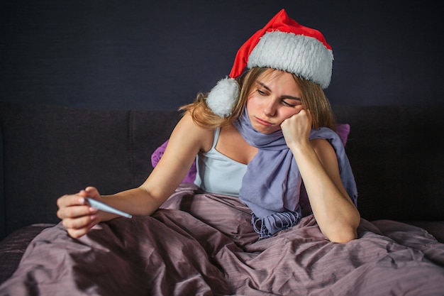 Sad and upset young woman sits on bed and looks on thermometer she holds in hand. Model holds Christmas hat on head. Young woman is bored.