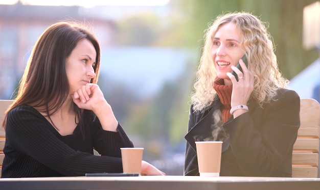 Foto donna triste ignorata dalla sua amica seduta al caffè di strada all'aperto mentre parla felicemente al telefono cellulare e non presta attenzione concetto di problemi di amicizia