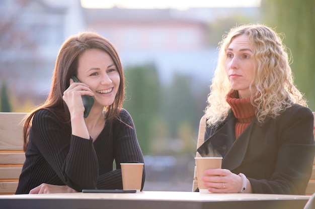 Foto donna triste ignorata dalla sua amica seduta al caffè di strada all'aperto mentre parla felicemente al telefono cellulare e non presta attenzione concetto di problemi di amicizia