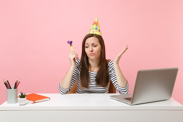 Photo sad woman in birthday hat with playing pipe feeling unhappy because nobody came to celebrate sit work at white desk with pc laptop isolated on pink background. achievement business career. copy space.