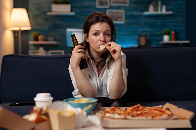 Sad woman drinking craft beer and eating potato chips from bowl at table with large pizza and takeout dinner in home living room. Tired office worker on couch relaxing after takeaway menu.