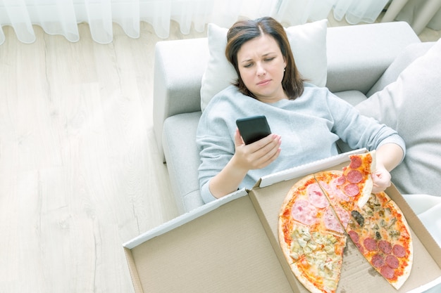 Donna triste che mangia pizza e che tiene telefono che mette su sofà a casa, tono blu