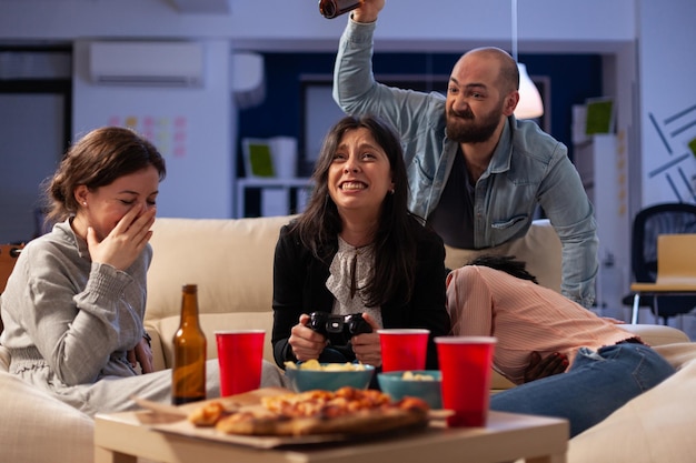 Sad woman losing video games play on tv console with controller, attending office party after work hours. Colleagues using online game play at drinks celebration with food snacks.