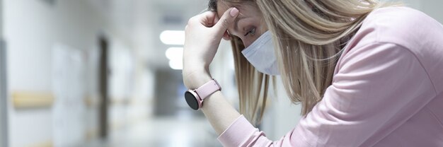 Грустная женщина в медицинской защитной маске сидит в коридоре клиники