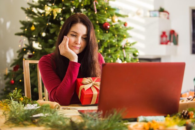 悲しい女性は彼女のラップトップコンピューターの画面でクリスマスのお祝いをオンラインで見ている彼女の家族を逃します