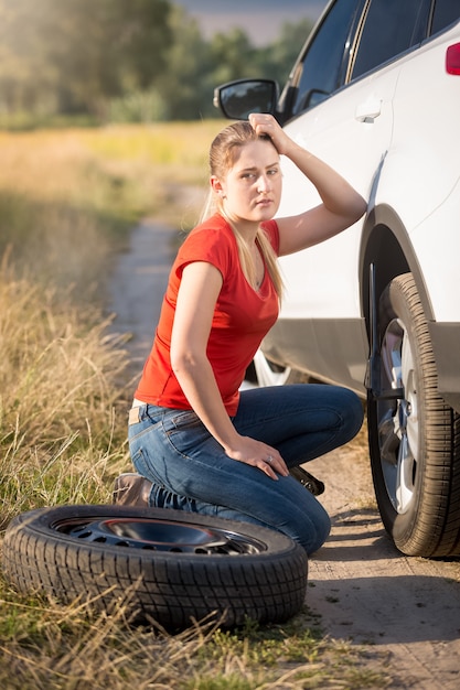 Sad woman sitting at broken car and trying to change flat tire