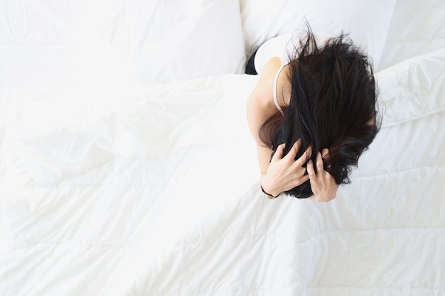 하얀 침대에 앉아 머리를 들고 있는 슬픈 여자 불면증 치료 개념