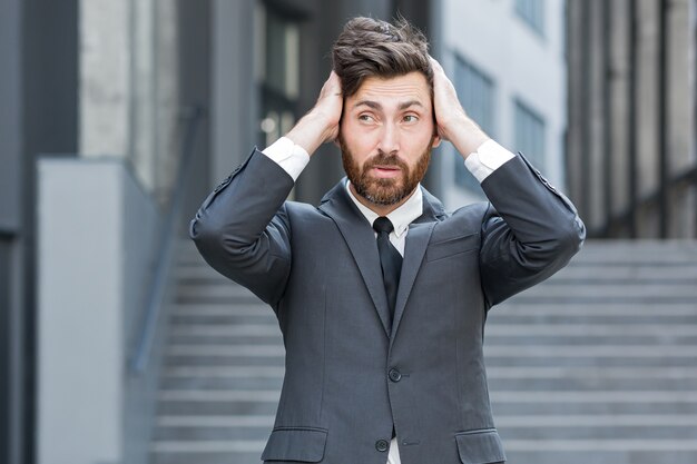 彼の頭を保持している悲しい心配ビジネスひげを生やした白人男性。現代のオフィスセンターを背景に街の通りでスーツを着たビジネスマンの起業家または従業員または労働者。外。屋外