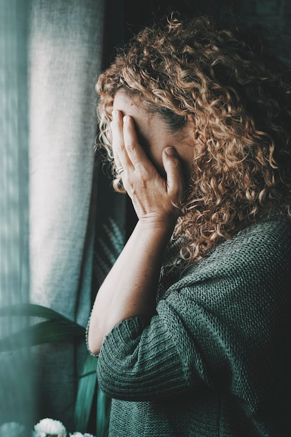 Sad and worried woman side portrait at home in the dark looking outside the window Worried and lonely female alone indoor Concept of end of relationship and unsuccessful emotion Lady touching face