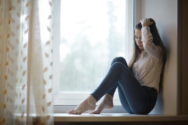 窓の上に座っている悲しい若い大人の女性