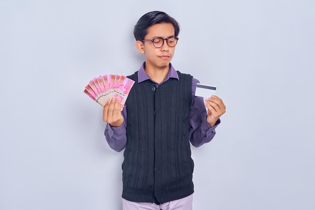 Sad young Asian man in vest shirt showing money rupiah banknotes and credit card isolated on white background