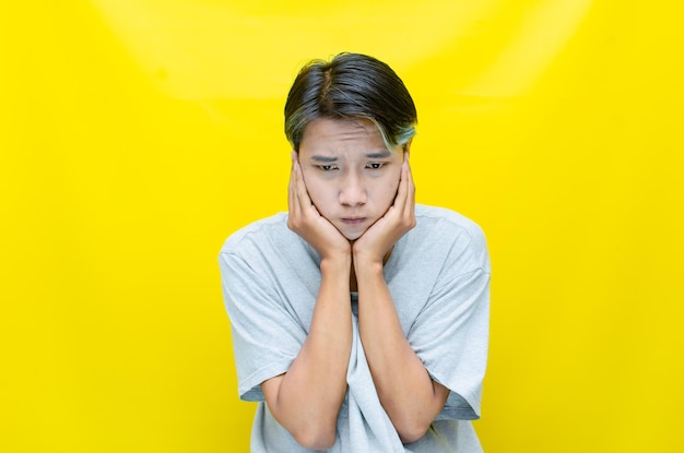 Sad young asian man wearing grey tshirt with crying face expression isolated over yellow background