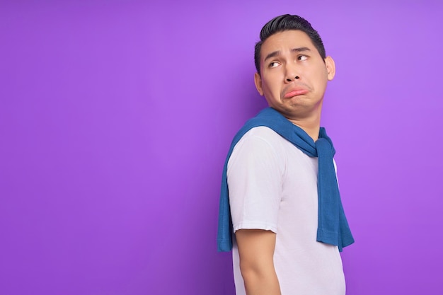 Sad young Asian man wearing white tshirt is standing and looking aside at copy space isolated over purple background people lifestyle concept