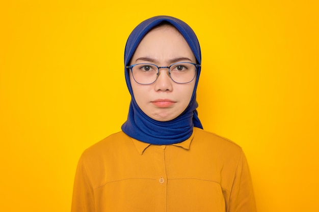 Sad young Asian Muslim woman dressed in orange shirt looking at camera with dissatisfied sadness face isolated on yellow background