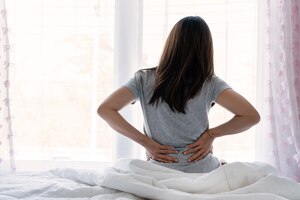 Sad young asian woman touching back feeling backache morning discomfort low lumbar muscular kidney pain sit on bed after bad sleep waking up on uncomfortable mattress bending. concept of woman stretch