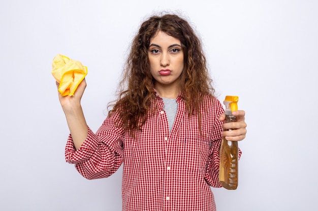 Sad young cleaning woman holding cleaning agent with rag isolated on white wall