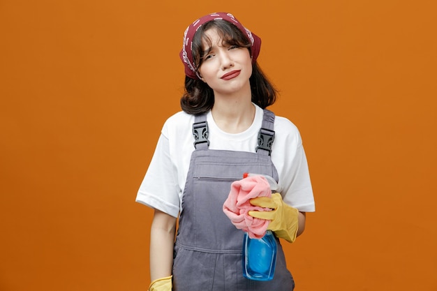 Sad young female cleaner wearing uniform rubber gloves and bandana holding cloth duster and cleanser looking at camera isolated on orange background