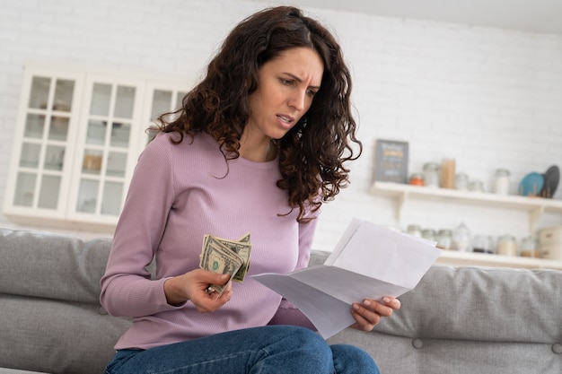 Sad young girl holding last cash money feeling anxiety about debt or bankruptcy sitting at home