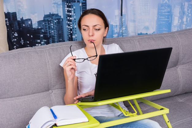 La ragazza triste ha messo gli occhiali alla bocca mentre lavorava su un laptop facendo i suoi affari da casa seduta su un divano nel soggiorno in un'atmosfera rilassata