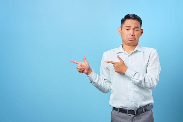 Sad young handsome businessman pointing finger away at copy space on blue background