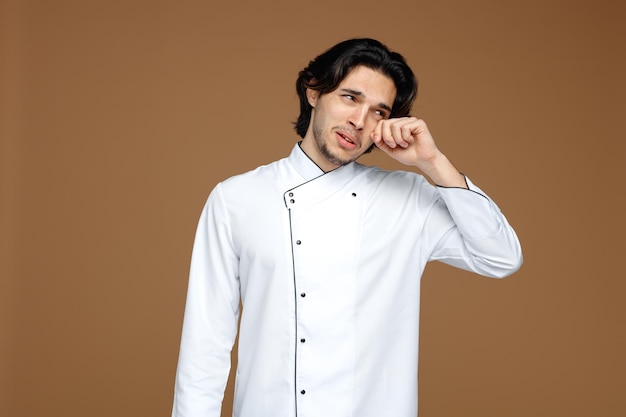 sad young male chef wearing uniform looking at side crying wiping eye with hand isolated on brown background