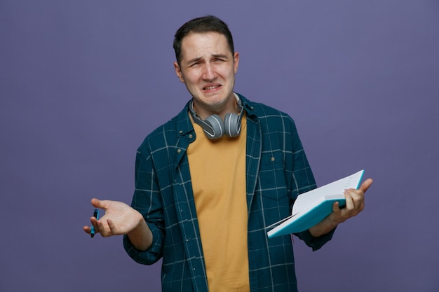 sad young male student wearing headphones around neck holding pen and note book looking at camera crying isolated on purple background