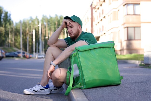 Foto un giovane corriere triste in uniforme verde con una grande borsa termica o uno zaino che consegna cibo dal ristorante