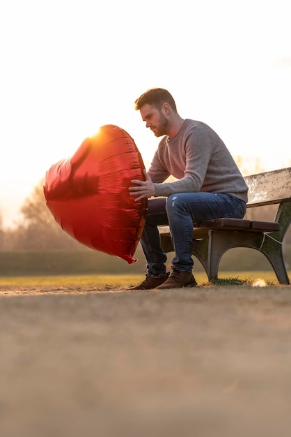 Sad young man holding up a heartshaped balloon sitting on a bench in the park and looking at the balloon