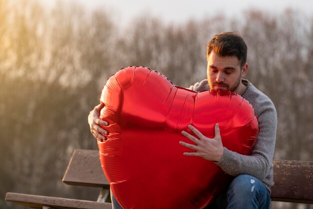 Photo sad young man hugging a heartshaped balloon sitting on a bench in the park and thinking with his eyes closed