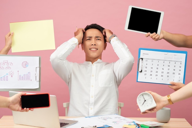 Photo sad young man sits at desktop hands with papers alarm clock touchpad notepad with stickers studies documentation isolated over pink background