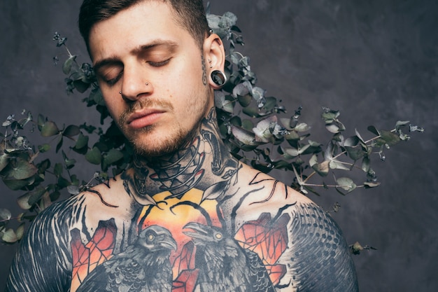 Sad young man with tattoo on his body standing in front of dry leaves against grey background