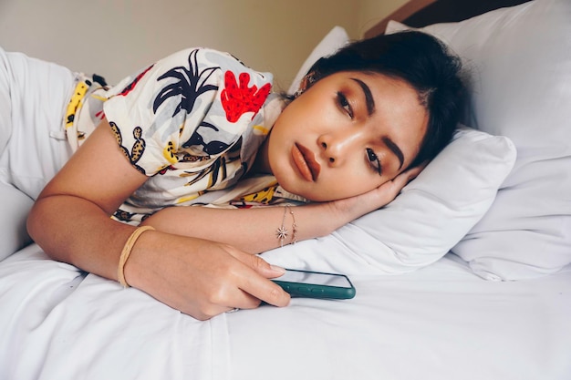 Грустная молодая женщина хмурится с кровати и ждет звонка, держа смартфон