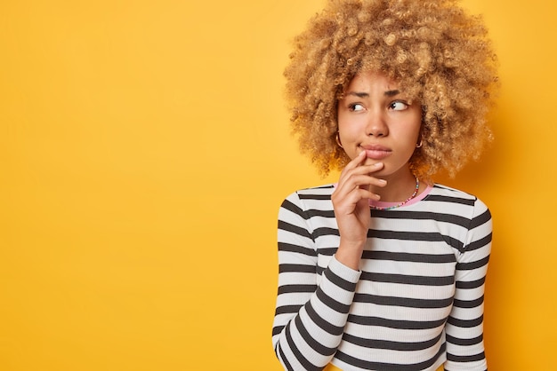 Sad young woman has thoughtful grumpy face being upset by something concentrated away wears casual striped jumper isolated over yellow background empty space for your promotion Let me think