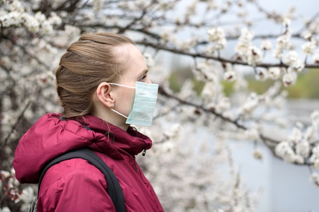 Sad young woman in protective medical face mask. Blooming tree on . Spring allergy.