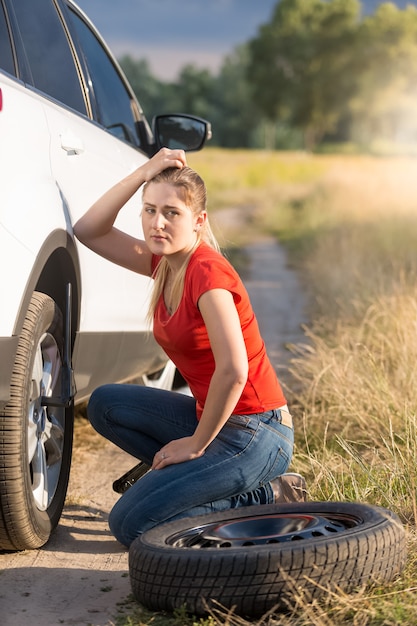 フィールドで車のパンクしたタイヤを交換する悲しい若い女性