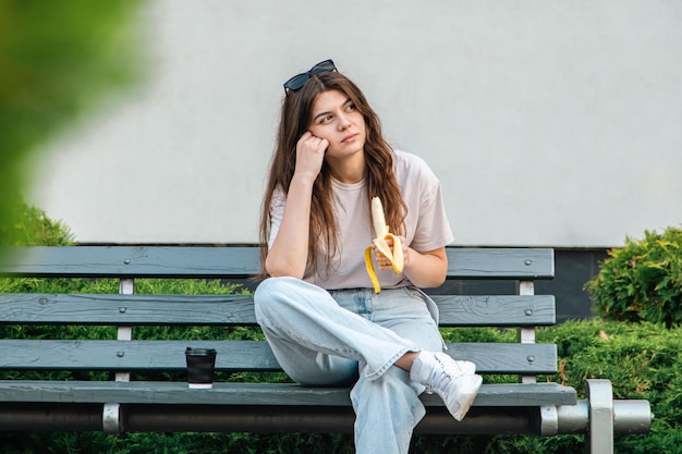 A sad young woman sits on a bench and eats a banana