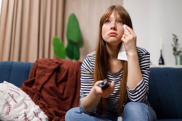 Sad young woman sitting on sofa with tissue paper and remote control in hand watching drama movie