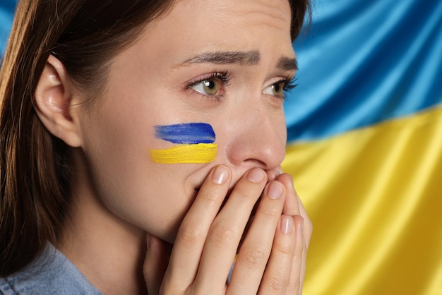 Sad young woman with clasped hands near Ukrainian flag closeup