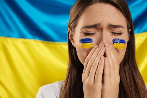 Sad young woman with clasped hands near Ukrainian flag Space for text