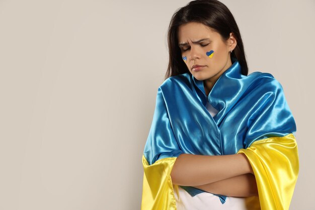 Sad young woman with face paint and Ukrainian flag on beige background space for text