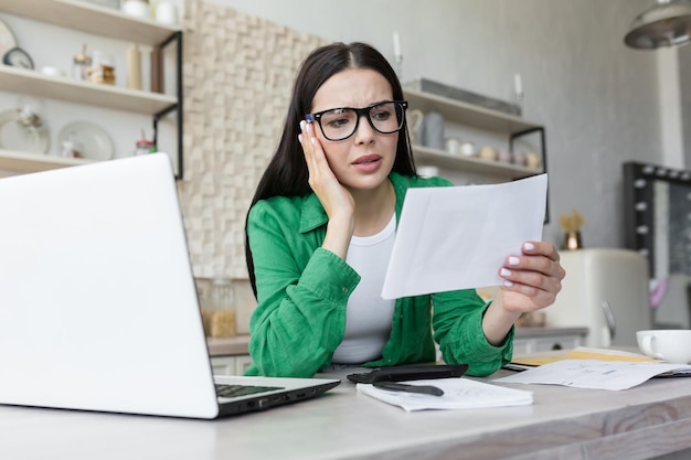 Sad young woman working from home with laptop and documents holds in hands and anxiously looks at