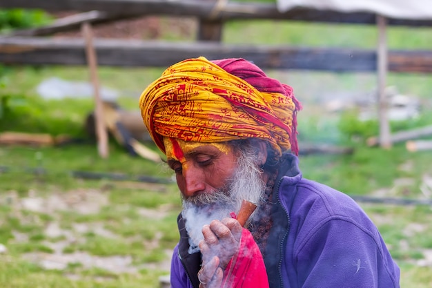 Sadhu man with traditional painted face smoking a pipe with special herbs for meditation