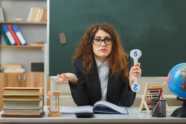 sadly spreading hand young female teacher wearing glasses holding number fan sitting at desk with school tools in classroom