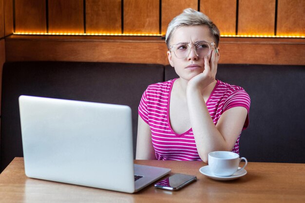 Sadness tired young girl freelancer with short hair in pink tshirt is sitting alone in cafe and searching for a work on laptop in bad mood without good result and thinking