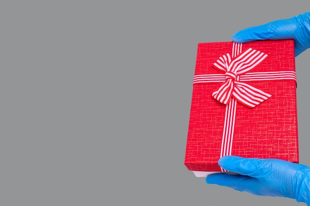 Safe delivery of gifts concept. Hands in blue gloves hold a red gift box with a bow on the trendy Ultimate Gray background. Safe gifts during the 2021 coronavirus pandemic