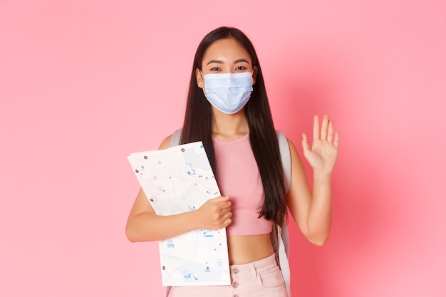 Safe tourism, Traveling during coronavirus pandemic and preventing virus concept. Friendly cute asian girl tourist with map and bacpack going abroad, waving hand hello, wear medical mask