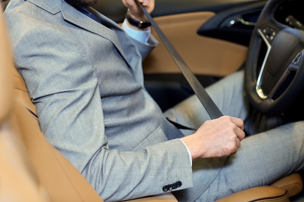 safety, driving and people concept - close up of man in elegant business suit fastening seat safety belt in car