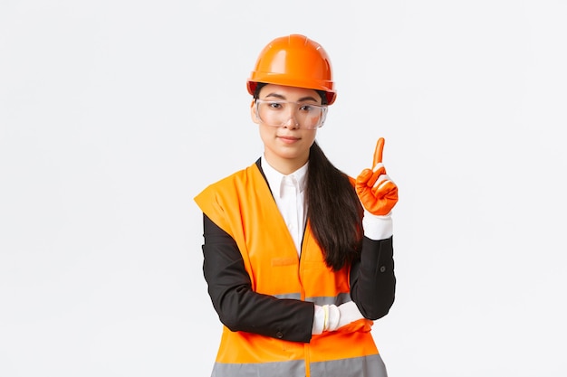 Safety first. Serious, smiling female asian construction engineer, industrial woman showing finger at telling rules on enterprise, wearing protective glasses, helmet and gloves, white background