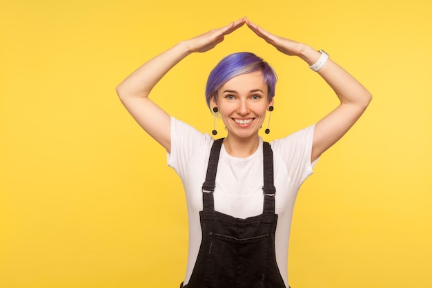 安全性。デニムのオーバーオールに紫のショートヘアを着た陽気なヒップスターの女の子の肖像画は、頭上に手を置いて屋根のシンボルを作り、笑顔で、家を夢見て、生命を確保しました。黄色の背景、スタジオショット