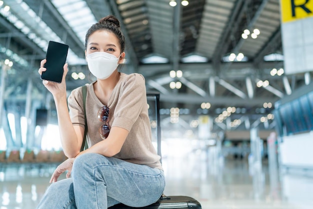 Safty travelyoung attraente donna asiatica donna indossare maschera viso gesto della mano mostra schermo vuoto smartphone viaggio vacanza dopo il blocco è donna overasiatica sedersi rilassarsi al terminal dell'aeroporto